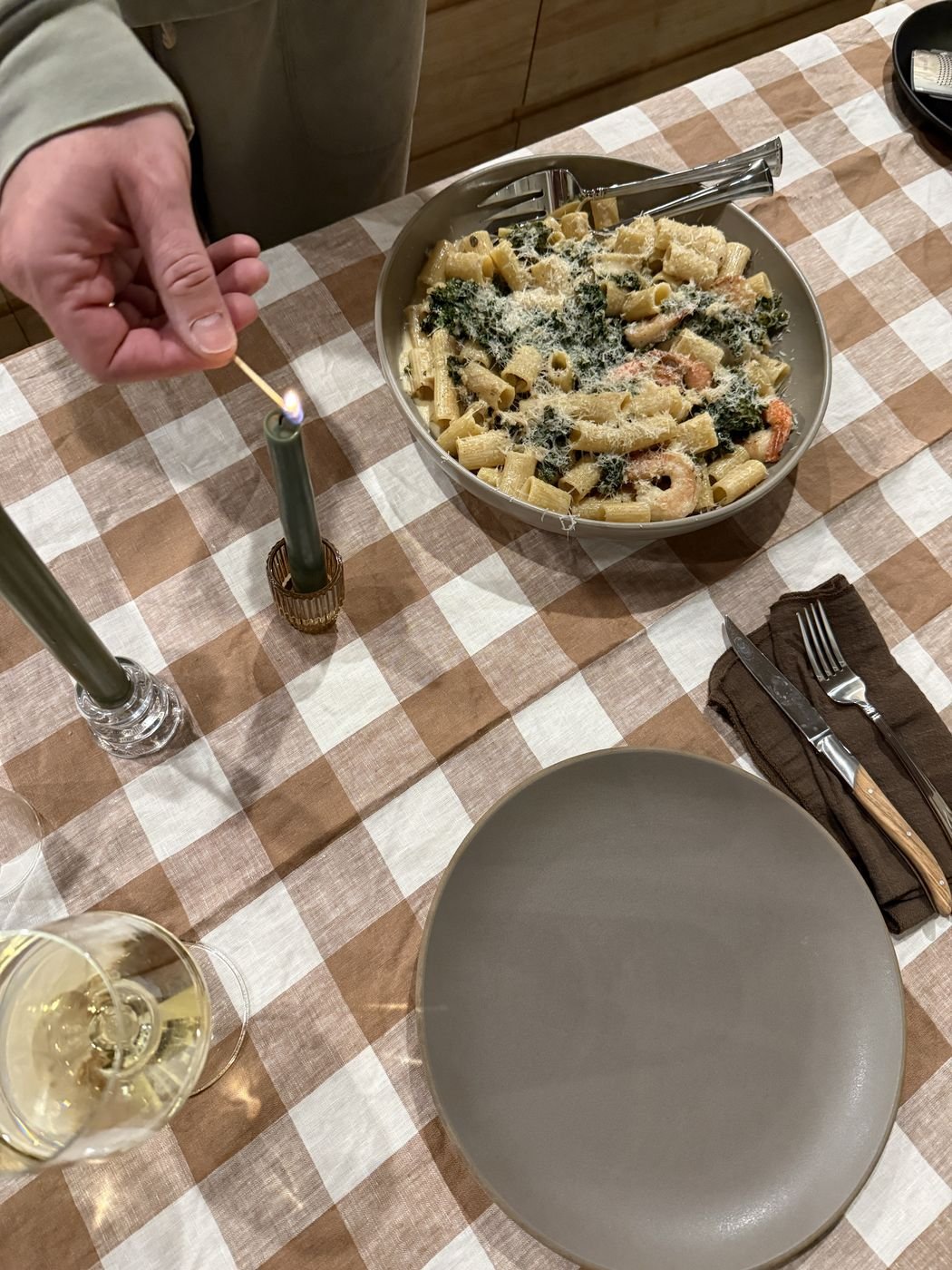 Lighting a candle at a gingham-covered table set with a bowl of pasta and white wine
